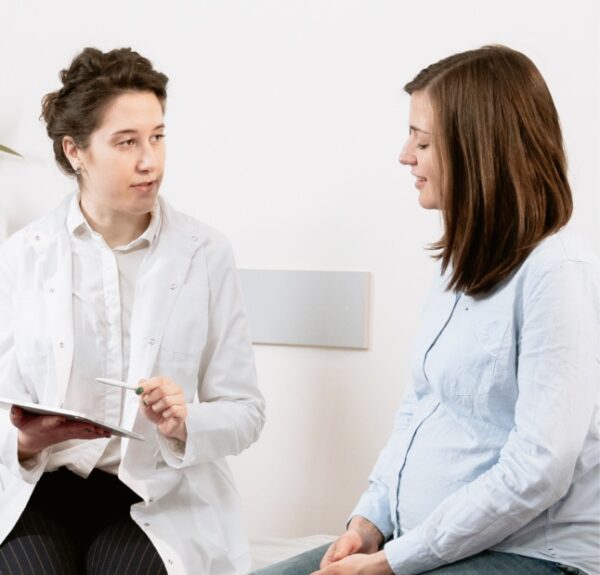 A female doctor holds a clipboard and speaks with a pregnant patient during a consultation.