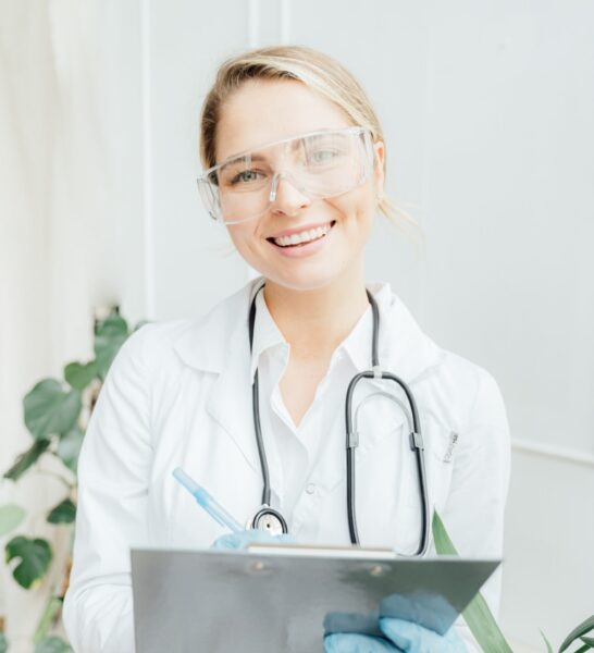 A smiling female doctor wearing safety goggles, a lab coat, and a stethoscope holds a clipboard.