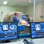 A radiology control room with computer monitors displaying chest scans, looking through a window at a patient in a CT scanner.