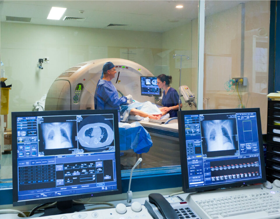 A radiology control room with computer monitors displaying chest scans, looking through a window at a patient in a CT scanner.
